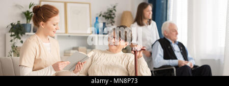 Krankenschwester Lesung eine ältere Frau mit einem Stock und ein älterer Mann im Rollstuhl von einem Freiwilligen in einem Pflegeheim Pflegeheim geschoben Stockfoto