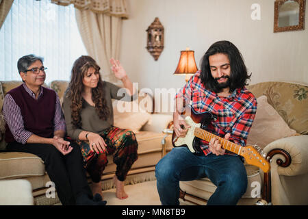 Mitte der erwachsenen Mann spielt Gitarre, während seine Eltern nach Hause zu Besuch. Sie sind auf dem Sofa sitzen, Tanzen und Singen im Hintergrund. Stockfoto