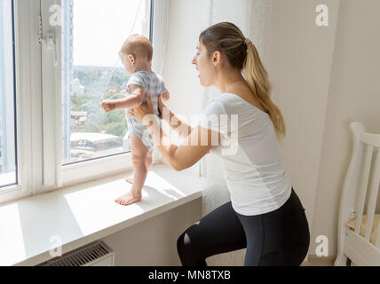 Junge Mutter fangen und halten Ihr Baby Sohn stehen auf sindowsil und Suchen aus dem Fenster Stockfoto
