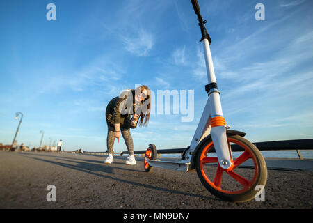 Attraktive Mädchen und ihre Roller auf dem Asphalt. Junges Küken lächelnd und Sieg Symbol angezeigt. Weiß kick Scooter mit orange Räder. Sportliche Aktivität. Stockfoto