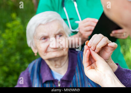 Ältere kranke Frau, die Pillen von Krankenschwester outdoor, Arzt schreiben ärztliche Verschreibung im Hintergrund Stockfoto