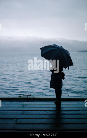 Frau mit Blick über den Loch Lomond mit einem Regenschirm auf einem nebligen kalten Wintern. Stockfoto