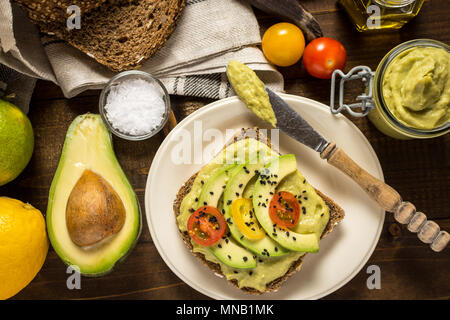 Verbreiten frischen Avocado Guacamole und Scheiben mit Cherry Tomaten und schwarzem Sesam auf ein Stück Brot. Gesundes Frühstück Konzept. Stockfoto