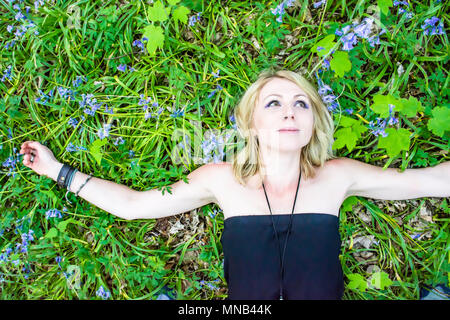 Junge, blonde Frau liegend auf Waldboden voller Bluebells, die Arme weit ausgebreitet und Lächeln auf ihrem Gesicht. POV Bild. Positive Zukunft. Stockfoto