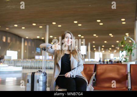 Gerne Frau sitzt im Flughafen Halle mit modernen Tablet in der Nähe von valise und Daumen hoch. Stockfoto