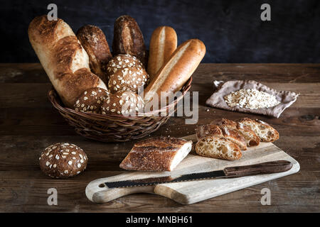 Eine Auswahl an Brot in einem Korb und ein Baguette auf dem Schneidebrett. Stockfoto