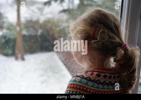 Kleines Mädchen mit Blick durch das Fenster auf den verschneiten Garten in Cornwall, England Stockfoto