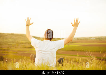 Mann sitzt auf dem Feld und Anheben der Arme bis Stockfoto