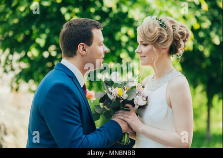 Glücklich lächelnde stilvolle Braut und Bräutigam im Sommer, grünen Park mit Blumenstrauß, Tanzen und Spaß am Tag ihrer Hochzeit Stockfoto