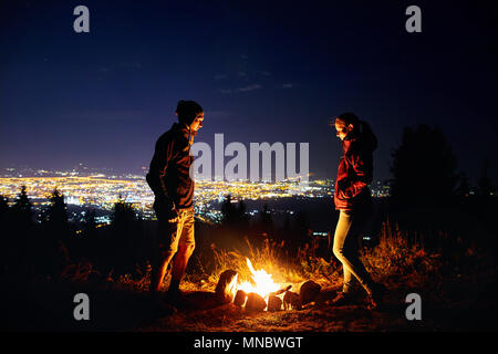 Glückliches Paar Wanderer wärmen Sie die Hände in der Nähe von Lagerfeuer unter Sternenhimmel mit Sterne und die Lichter der Stadt im Hintergrund. Stockfoto