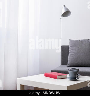 Buch und zwei Tassen liegen auf einem kleinen Tisch. Zimmer mit weißen Vorhang, Lampe und graue Sofa. Stockfoto