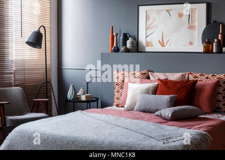 Kreative Poster auf Schwarz bedhead über gemütliches Bett mit dekorativen Kissen und graue Decke im Schlafzimmer Innenraum mit Metall Lampe und Vintage Sessel. Re Stockfoto