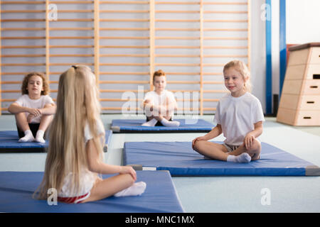 Glückliche junge Mädchen sitzen einander gegenüber auf blauen Matten Trainieren mit ihren Beinen während PE-Klassen in der Grundschule Turnhalle Innenraum gekreuzt Stockfoto