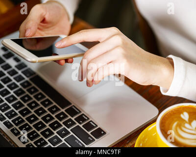 Nahaufnahmen der Hände einer Frau mit Handy und Laptop beim trinken Cappuccino. Stockfoto