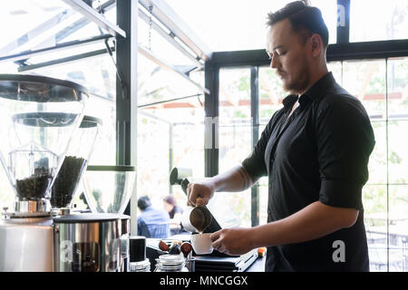 Ernsthafte Barkeeper gießen frische Milch in eine Tasse Kaffee Stockfoto