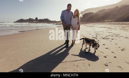 Volle Länge geschossen von Senior Paar ihren Hund am Strand entlang am Morgen. Reifes Paar auf dem morgendlichen Spaziergang mit ihrem Hund. Stockfoto