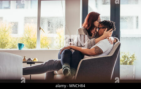 Frau küssen Freund Stirn mit Liebe, während auf einem Stuhl sitzen zusammen. Romantische interracial Paar zu Hause. Stockfoto