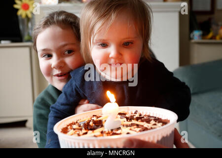 Geburtstag Überraschung, eine Nummer zwei Kerze von den Geburtstag Mädchen mit ihrer Schwester, die ein freches Lächeln geblasen wird. England. Vom 7. März 2018 Stockfoto