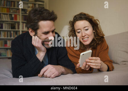 Lächelnde Frau sharing smart phone mit der Mann auf dem Bett zu Hause liegen Stockfoto