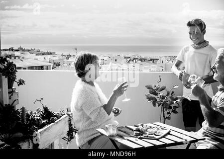 Großväter und Neffe blonde junge Aufenthalt auf der Terrasse trinken und essen Essen auf den Tisch. Tolle Aussicht aufs Meer und klaren Himmel für Schwarz und Weiß Ich Stockfoto