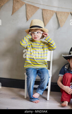 Happy Boy trug Hut und Brille sitzen auf Stuhl von Freund gegen die Wand Stockfoto