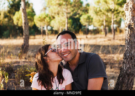 Mädchen küssen glücklich Vater von Baumstamm sitzen im Wald Stockfoto