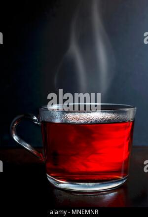 Hot red berry Obst Kaffee in einem Glas Becher mit Dampf stehend auf einem dunklen Holz mit dunkelgrauer Hintergrund