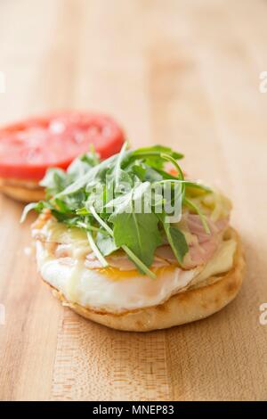 Ein englischer Muffin mit Eiern, Käse, Schinken Rucola und Tomaten zum Frühstück Stockfoto