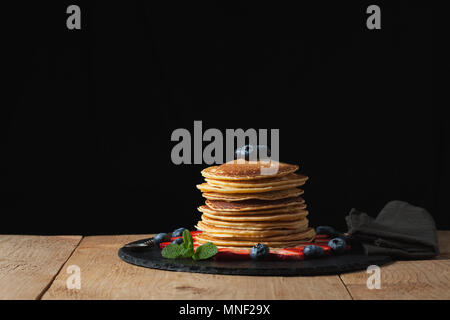 Vorderansicht des Stack von hausgemachten plain Pfannkuchen mit Erdbeeren, Heidelbeeren und Ahornsirup auf schwarzem Teller serviert auf schwarzen Hintergrund mit selektiven Fokus. Mit Platz kopieren Stockfoto