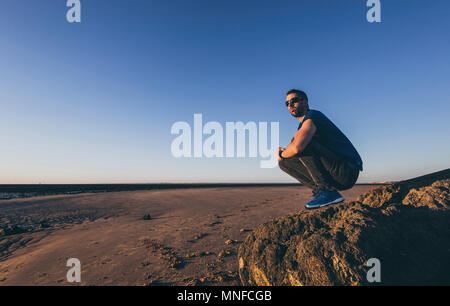 Junger Mann kauert auf einem Felsen mit Sonnenbrille am Strand von Sanlucar de Barrameda Stockfoto