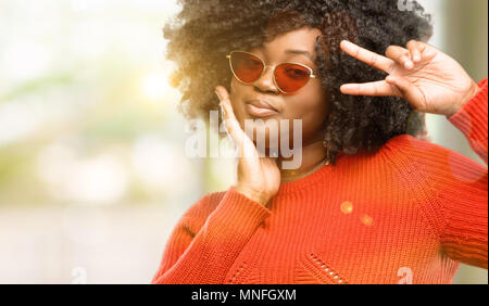 Schöne afrikanische Frau an der Kamera durch ihre Finger im Sieg Geste zwinkerndes Auge und bläst einen Kuss, outdoor Stockfoto