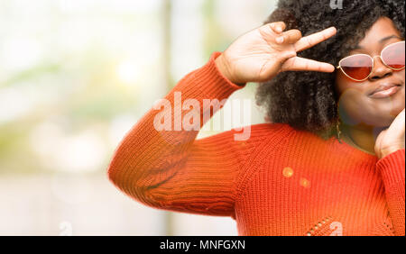 Schöne afrikanische Frau an der Kamera durch ihre Finger im Sieg Geste zwinkerndes Auge und bläst einen Kuss, outdoor Stockfoto