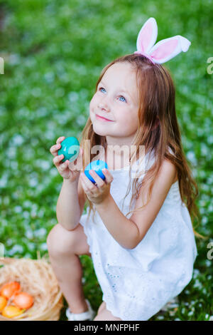 Adorable kleine Mädchen mit Hasenohren spielen mit Ostereier im Gras mit weißen Blütenblätter auf Frühling abgedeckt Stockfoto