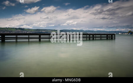 Hölzerne Seebrücke am Zürichsee mit sanften Hügeln Berglandschaft und Segelboote im Hintergrund Stockfoto