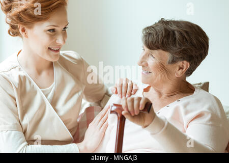 Elder glücklich Frau mit hölzernen Spazierstock und Gespräch mit ihr junge Krankenschwester Stockfoto
