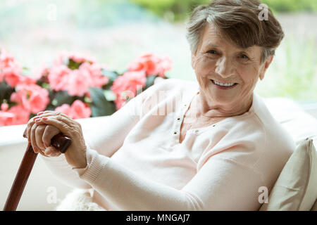 Ältere Dame sitzen auf der Couch mit hölzernen Spazierstock und lächelnd Stockfoto