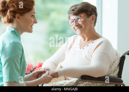 Junge kaukasier Krankenschwester unterstützt ältere Frau mit Brille im Rollstuhl Stockfoto