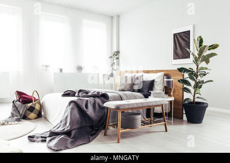 Holzbank und Anlage neben dem Bett mit grau Bettwäsche in hellen Schlafzimmer Innenbereich mit schwarzem Poster an der Wand Stockfoto