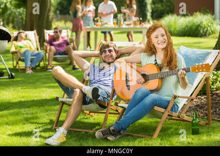 Frau sitzt auf der Sonnenbank, Gitarre spielen im Garten Stockfoto