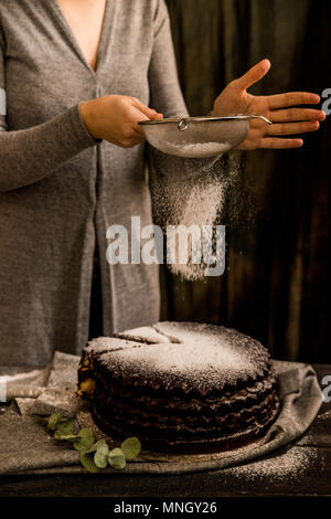 Close up Frau verzieren Schokolade Kuchen mit Zucker Pulver. Dark vintage Stilleben Stockfoto