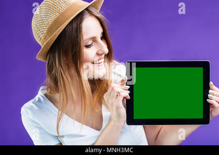 Lächelnde blonde Mädchen im weißen T-Shirt und Hut hält Tablet Computer mit Green Screen vor dem blauen Hintergrund, Raumkonzept werben Stockfoto