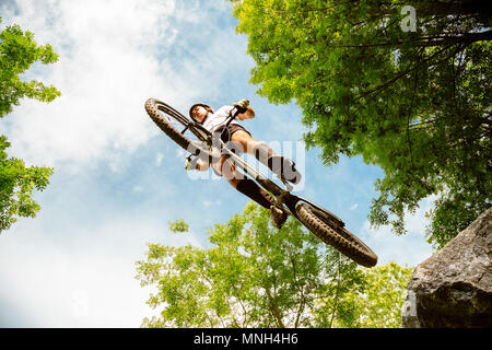 Junge Radfahrer fliegen mit seinem Fahrrad von einem Felsen im Wald. Extreme Low Angle View Stockfoto