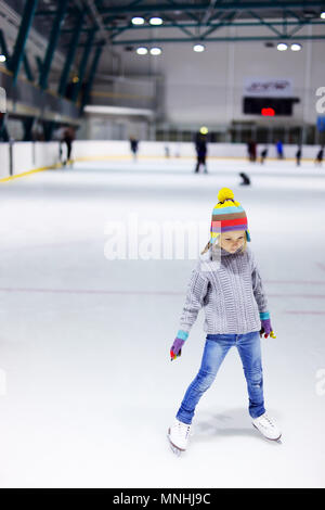 Adorable kleine Mädchen in Jeans, warmer Pullover und bunten Hut Schlittschuhlaufen auf der Eisbahn Stockfoto