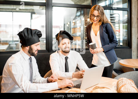 Mit zwei eleganten Bäcker oder Koch Köche arbeiten mit Laptop im Cafe mit Küche auf dem Hintergrund Geschäftsfrau Stockfoto