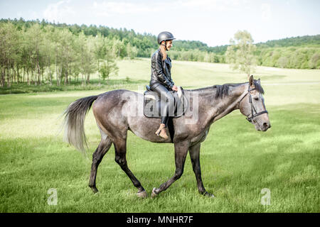 Frau in Leder Jacke mit Schutzhelm ein Pferd Reiten auf der grünen Wiese Stockfoto