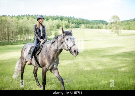 Frau in Leder Jacke mit Schutzhelm ein Pferd Reiten auf der grünen Wiese Stockfoto