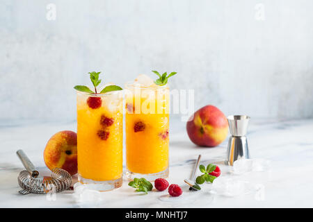 Zwei erfrischende Getränke von Pfirsich Saft und Himbeeren mit frischer Minze in hohe Gläser auf einem Marmortisch mit einem Shaker und bar Zubehör. Stockfoto