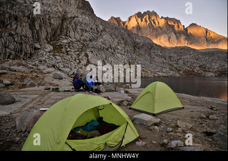 Backpackers Essen mit Scheinwerfern am Lager sitzen auf dem Barrett Seen in Palisade Becken auf einer zwei-wöchigen Trek der Sierra Hohe Weg in Kings Canyon National Park in Kalifornien. Die 200 km Route etwa Parallels die beliebte John Muir Trail durch die Sierra Nevada von Kalifornien von Kings Canyon National Park, Yosemite National Park. Sonnenuntergang leuchtet im Norden Palisade im Hintergrund. Stockfoto