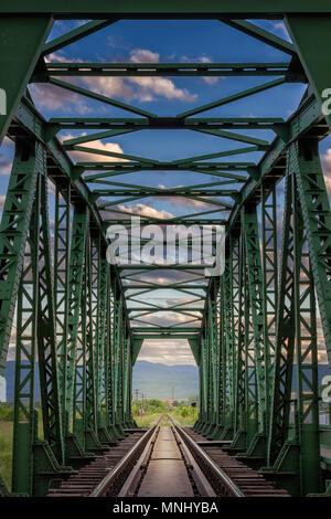 Vertikale Ansicht durch alte, Bügeleisen, Eisenbahnbrücke bei Sonnenuntergang mit dramatischen, bunten Wolken und andere sonnenbeschienenen Seite Stockfoto