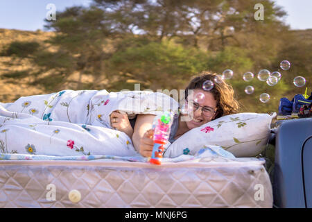 Frau mit einem Lächeln concentraited Spielen mit runden Seifenblasen im Bett, auf einen Pick up truck gebaut, während outdoor Reisen. Mit einem blury Baum Hintergrund Stockfoto